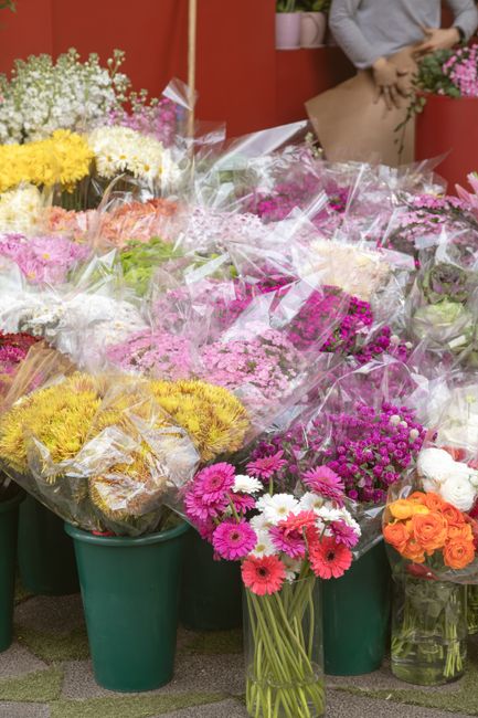 A display of freshly cut flowers arranged in green plastic buckets and vases at Crouch End Flowers, featuring vibrant pink gerbera daisies, orange roses, yellow chrysanthemums, and delicate purple and