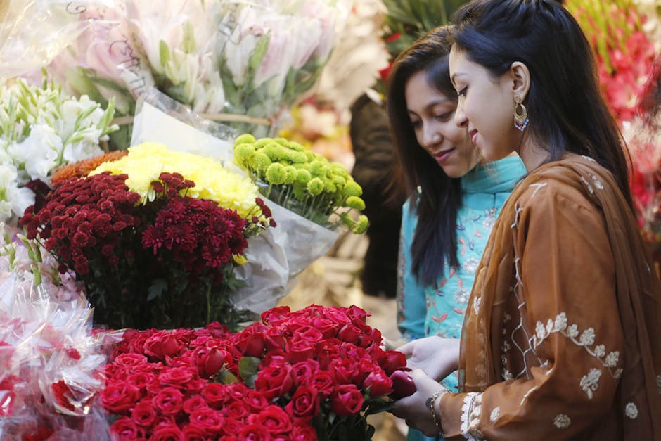 Two women are shopping for floral gifts at a flower shop, with various vibrant bouquets of fresh flowers displayed around them. The woman on the right, dressed in a brown embroidered jacket with white