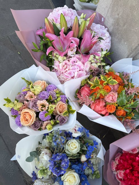 A collection of five vibrant floral arrangements, each wrapped in pastel-colored paper, displayed outdoors on a paved surface. The largest bouquet features pink lilies and pale pink hydrangeas, accent