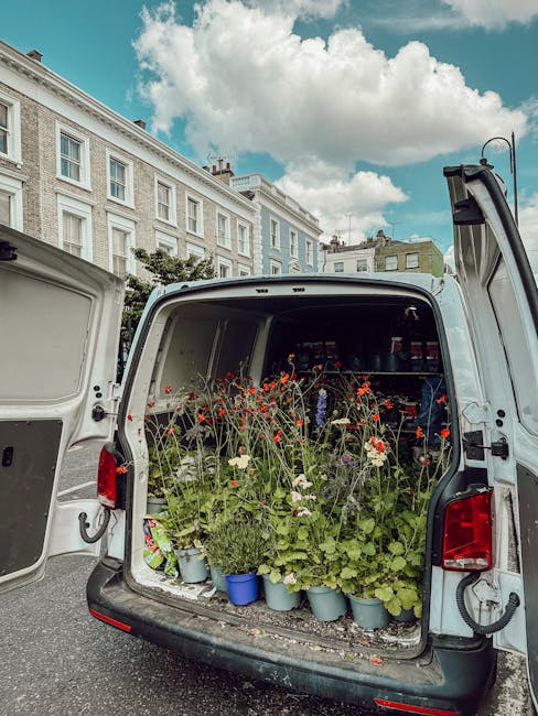 The open back of a white delivery van filled with potted flowers and plants, including red and white blooms, green foliage, and small decorative pots, parked on a street with a row of white terraced b