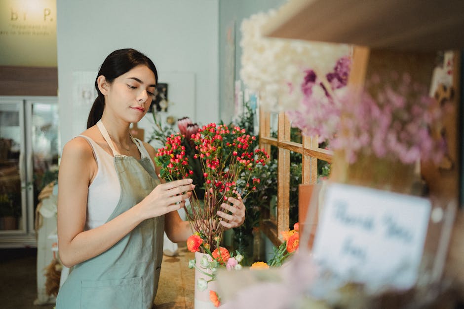 A young woman with dark hair tied back, dressed in a sleeveless white top and a light beige apron, is examining a bouquet of small pink waxflower blooms with green stems at a flower shop. The arrangem
