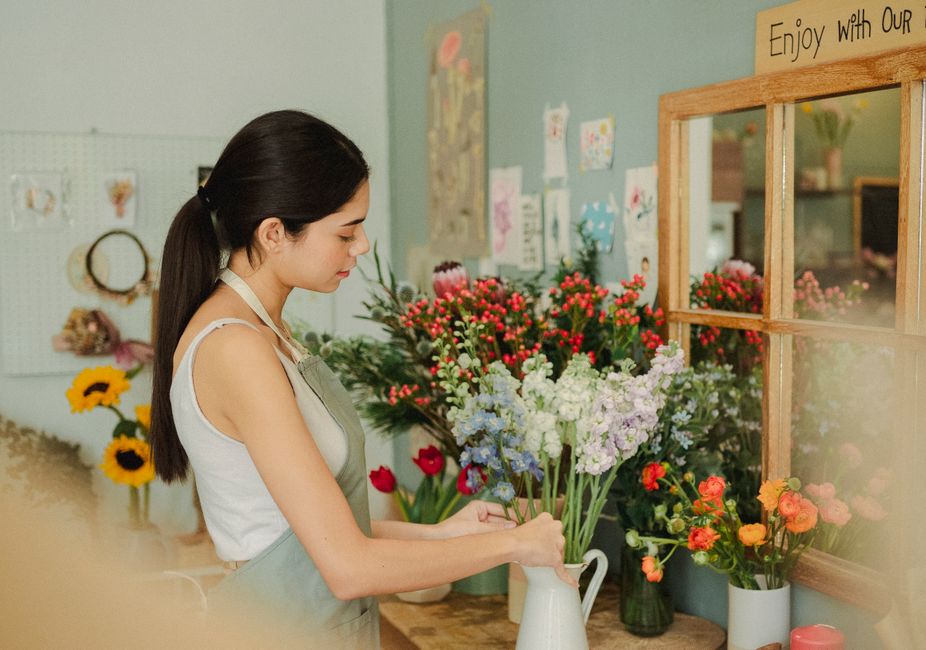 A young woman with long dark hair tied back related to a florist shop, wearing a white sleeveless top and a light green apron, arranging a bouquet of flowers in a white ceramic pitcher. The bouquet in